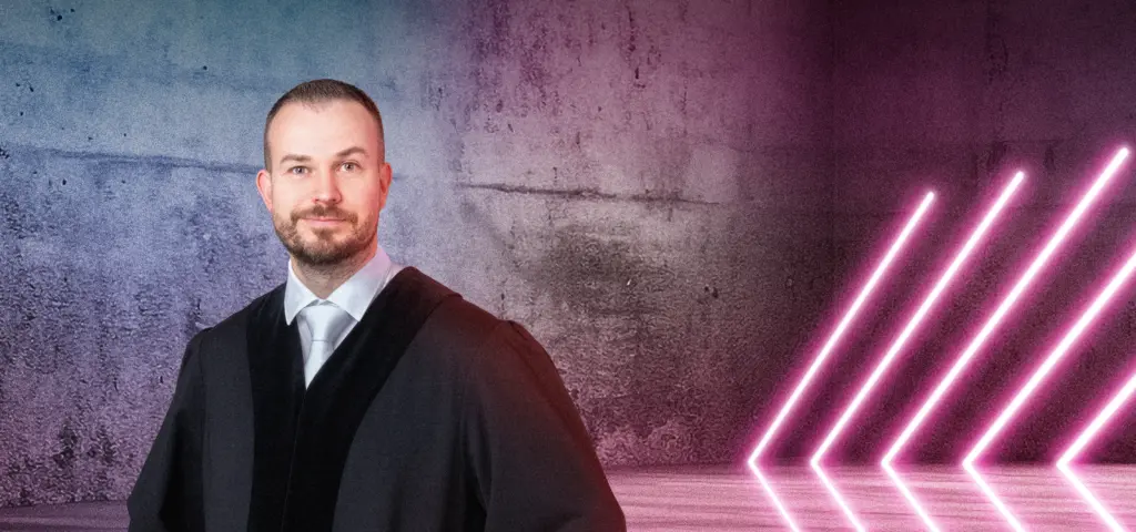 Eine Person in Robe steht lächelnd vor einer Betonwand mit blau-pinkem Lichtverlauf. Rechts im Bild leuchten mehrere diagonale Neonlichtstreifen. Die Szene wirkt modern und professionell.