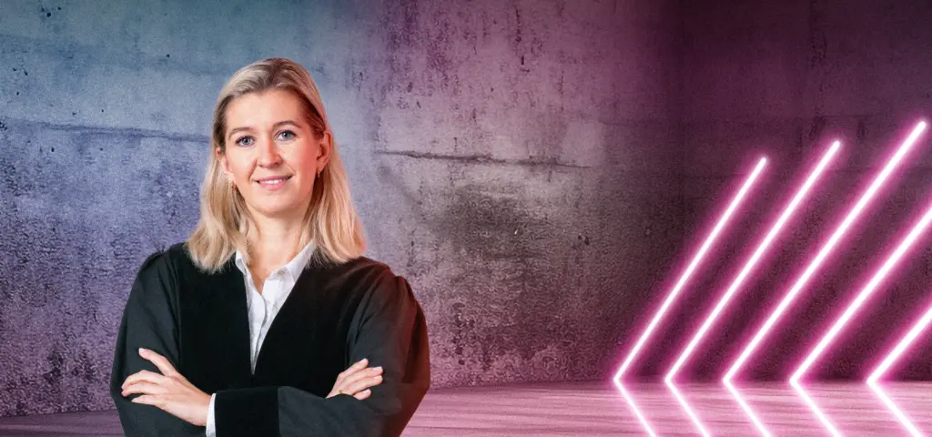 Eine Person in Robe steht lächelnd vor einer Betonwand mit blau-pinkem Lichtverlauf. Rechts im Bild leuchten mehrere diagonale Neonlichtstreifen. Die Szene wirkt modern und professionell.