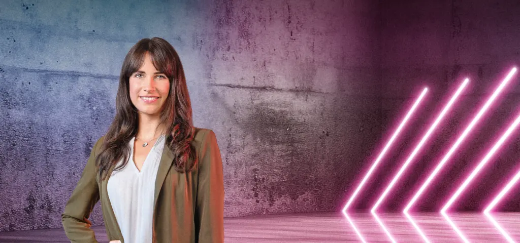 Eine Person steht lächelnd vor einer Betonwand mit blau-pinkem Lichtverlauf. Rechts im Bild leuchten mehrere diagonale Neonlichtstreifen. Die Szene wirkt modern und freundlich.