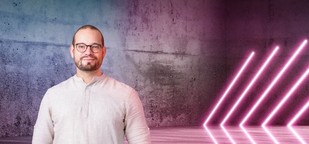 Eine Person steht lächelnd vor einer Betonwand mit blau-pinkem Lichtverlauf. Rechts im Bild leuchten mehrere diagonale Neonlichtstreifen. Die Szene wirkt modern und freundlich.