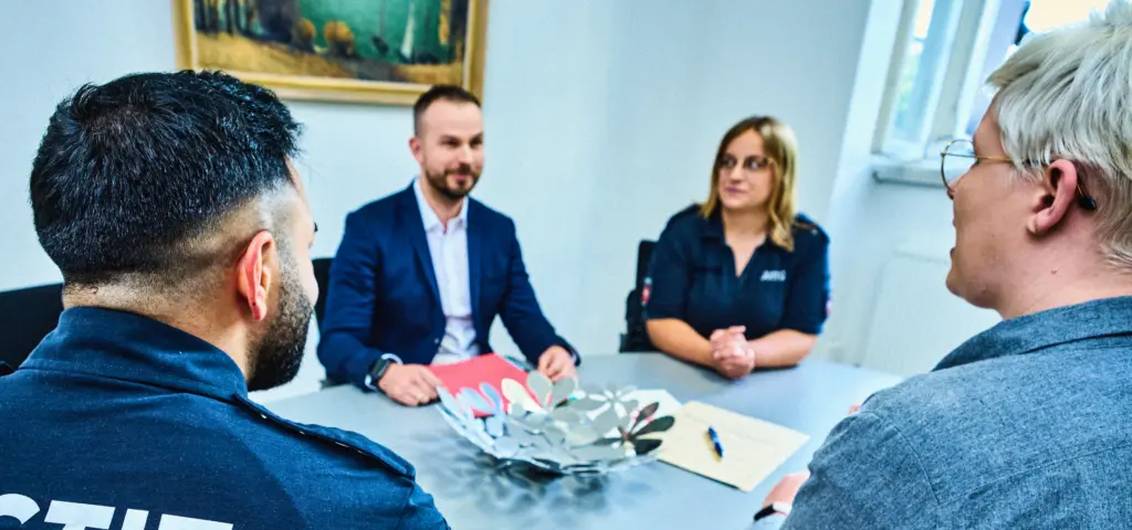 Vier Personen sitzen um einen Tisch in einem Besprechungsraum. Zwei Personen tragen Kleidung mit der Aufschrift „Justiz“, die anderen beiden formelle Kleidung. Auf dem Tisch liegen Unterlagen und ein Dekogefäß. Die Szene wirkt ruhig und konzentriert.