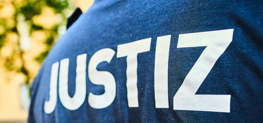 Nahaufnahme der Rückenpartie einer Person, auf deren dunklem Kleidungsstück groß das Wort „JUSTIZ“ steht. Der Hintergrund ist unscharf und zeigt helle Farben.