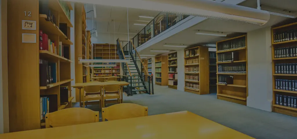 Ein heller Bibliotheksraum mit zahlreichen Bücherregalen, Tischen und Stühlen. In der Mitte führt eine Metalltreppe zu einer oberen Ebene mit weiteren Regalen. Die Atmosphäre wirkt ruhig und geordnet.