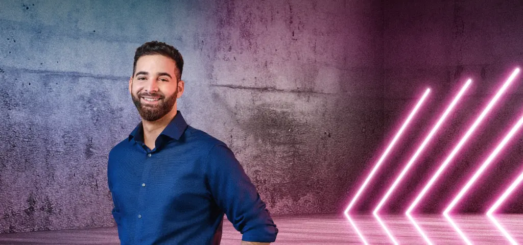 Eine Person steht lächelnd vor einer Betonwand mit blau-pinken Lichtverläufen. Rechts im Bild sind mehrere diagonale Neonlichtstreifen zu sehen, die in den Raum weisen. Die Szene wirkt modern und freundlich.