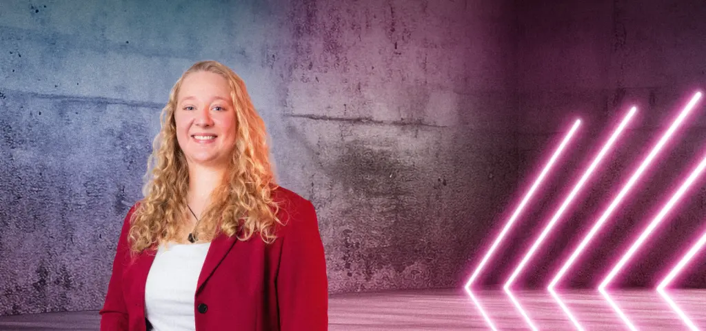 Eine Person steht lächelnd vor einer Betonwand mit farbigem Lichtverlauf in Blau und Pink. Rechts im Bild befinden sich mehrere diagonale Neonlichtstreifen, die in den Raum zeigen. Die Szene wirkt modern und farbintensiv.