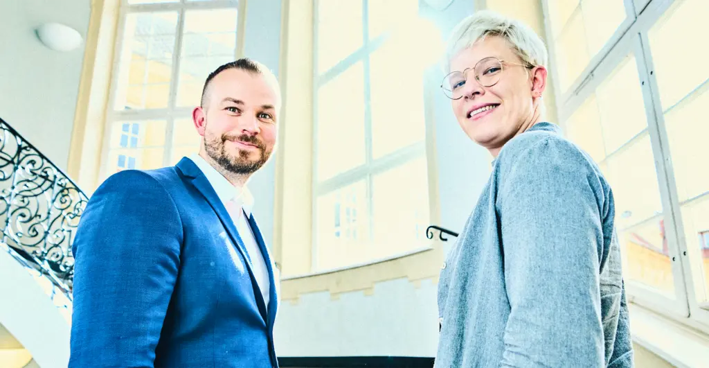 Zwei Personen stehen in einem hellen Treppenhaus vor großen Fenstern und blicken lächelnd zur Kamera. Das Geländer im Hintergrund ist verziert, und der Raum wirkt freundlich und lichtdurchflutet.