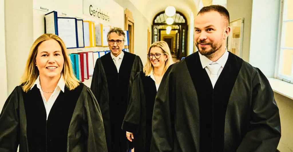 Vier Personen in Roben gehen gemeinsam durch einen hellen Flur eines Gerichtsgebäudes und lächeln. An der Wand hängen farbige Ordner, und im Hintergrund sind Rundbögen sowie Lampen zu sehen. Die Szene wirkt freundlich und professionell.