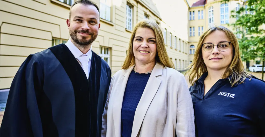 Drei Personen stehen im Außenbereich eines historischen Gebäudes und blicken lächelnd in die Kamera. Eine Person trägt eine Robe, eine andere Kleidung im Justizdienst, die dritte Person trägt eine helle Jacke. Im Hintergrund sind Gebäudefassaden und Bäume zu sehen.