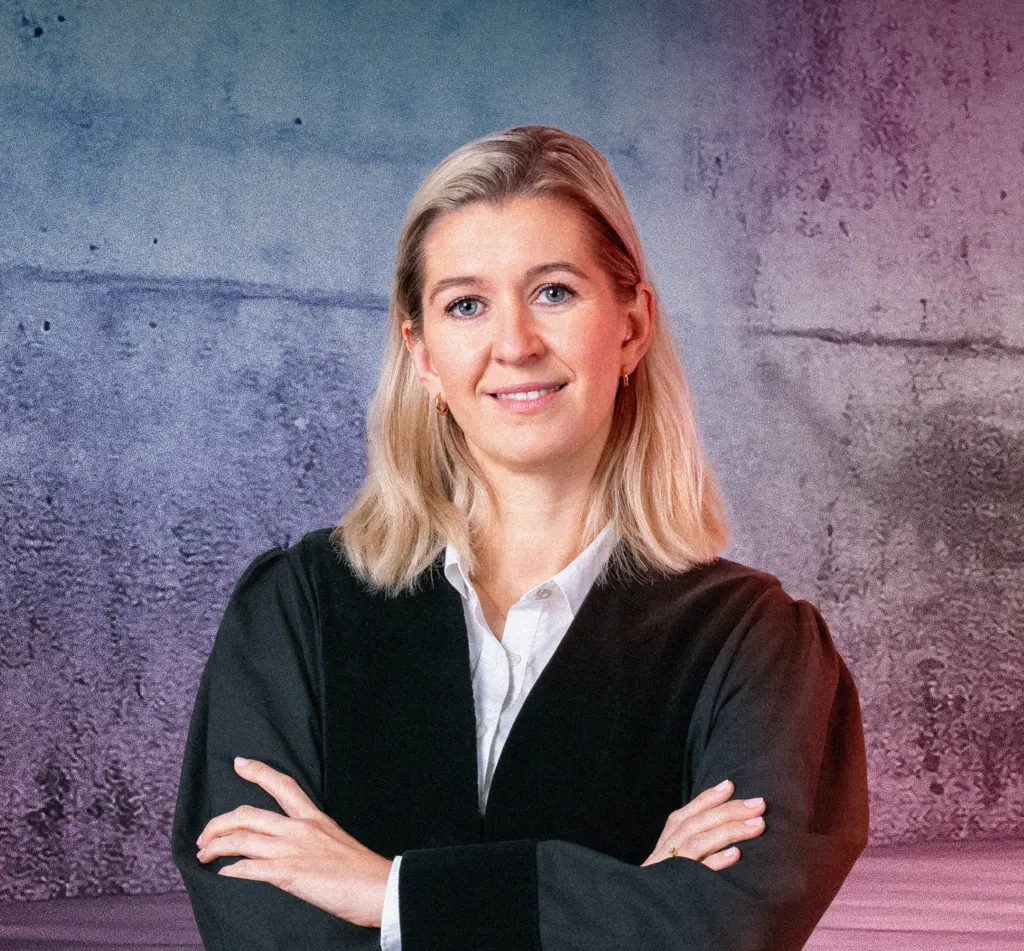 Eine Person in Robe steht lächelnd vor einer Betonwand mit blau-pinkem Lichtverlauf. Rechts im Bild leuchten mehrere diagonale Neonlichtstreifen. Die Szene wirkt modern und professionell.