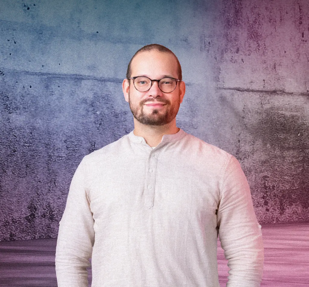 Eine Person steht lächelnd vor einer Betonwand mit blau-pinkem Lichtverlauf. Rechts im Bild leuchten mehrere diagonale Neonlichtstreifen. Die Szene wirkt modern und freundlich.