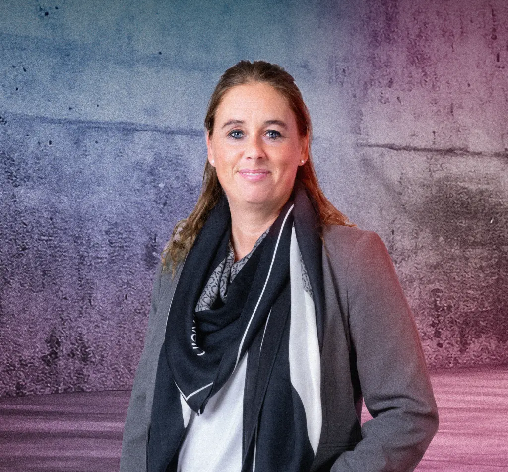 Eine Person steht lächelnd vor einer Betonwand mit blau-pinkem Lichtverlauf. Rechts im Bild leuchten mehrere diagonale Neonlichtstreifen. Die Szene wirkt modern und freundlich.