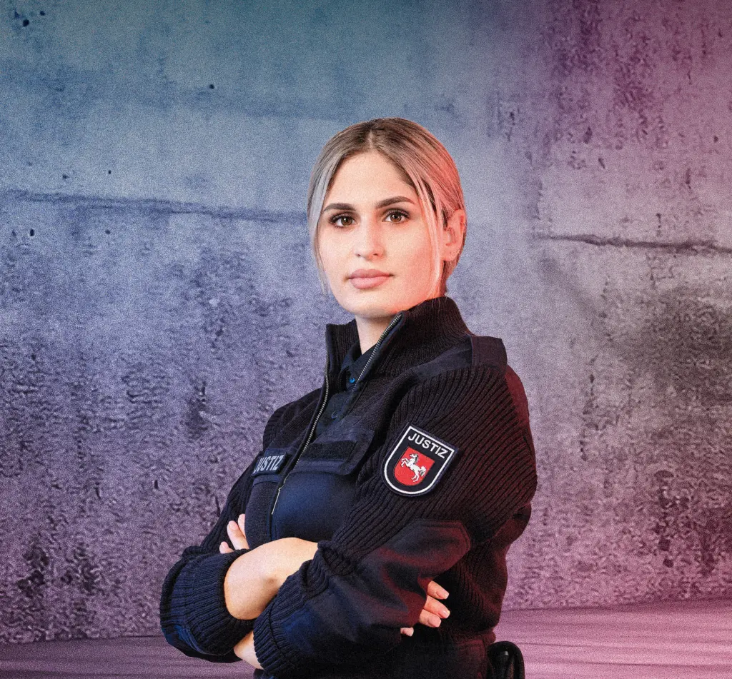 Eine Person in Justizdienstkleidung steht mit verschränkten Armen vor einer Betonwand mit blau-pinken Lichtverläufen. Rechts im Bild leuchten mehrere diagonale Neonlichtstreifen. Die Szene wirkt modern und selbstbewusst.