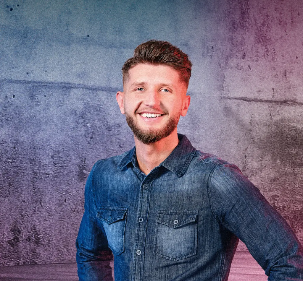 Eine Person steht lächelnd vor einer Betonwand mit blau-pinken Lichtverläufen. Rechts im Bild sind mehrere diagonale Neonlichtstreifen zu sehen, die in den Raum weisen. Die Szene wirkt modern und freundlich.