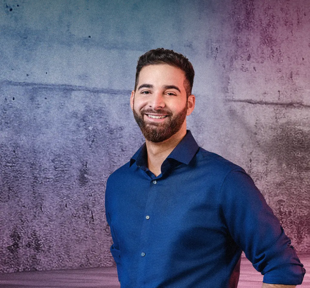 Eine Person steht lächelnd vor einer Betonwand mit blau-pinken Lichtverläufen. Rechts im Bild sind mehrere diagonale Neonlichtstreifen zu sehen, die in den Raum weisen. Die Szene wirkt modern und freundlich.