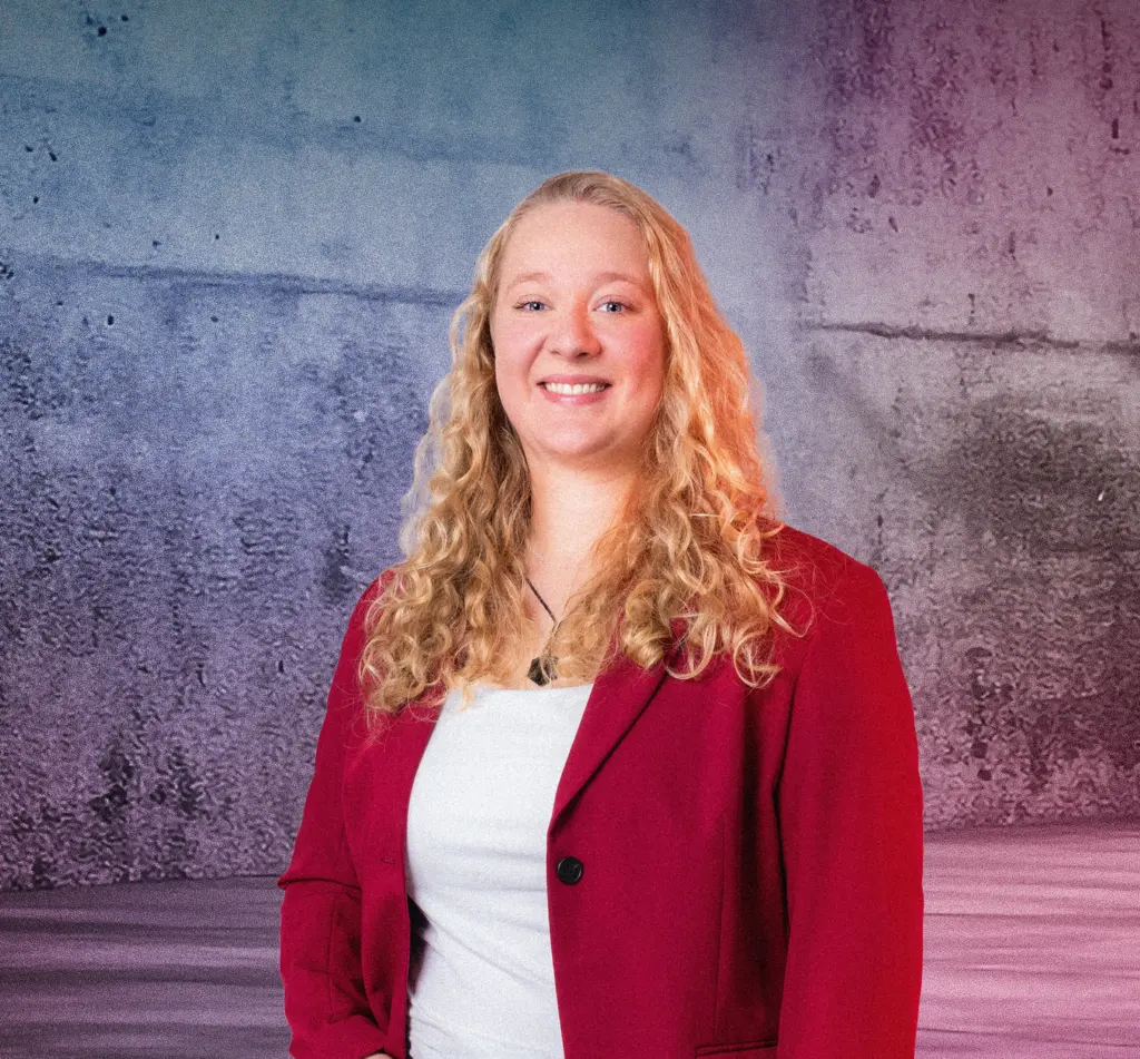 Eine Person steht lächelnd vor einer Betonwand mit farbigem Lichtverlauf in Blau und Pink. Rechts im Bild befinden sich mehrere diagonale Neonlichtstreifen, die in den Raum zeigen. Die Szene wirkt modern und farbintensiv.