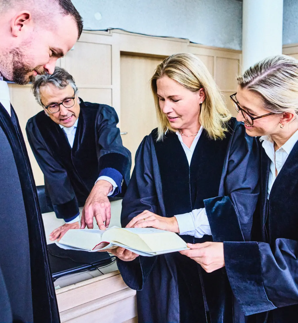 Vier Personen in Roben stehen zusammen und betrachten ein aufgeschlagenes Buch. Eine Person zeigt auf eine Stelle im Buch, während die anderen aufmerksam folgen. Die Szene wirkt konzentriert und kooperativ.