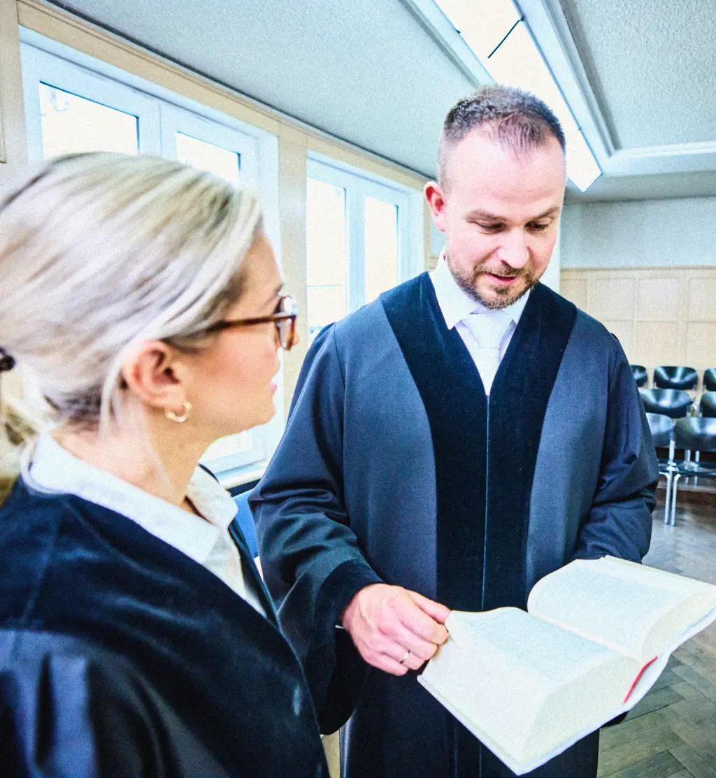 Zwei Personen in Roben stehen in einem hellen Raum und betrachten gemeinsam ein aufgeschlagenes Buch. Eine Person hält das Buch, die andere hört aufmerksam zu. Im Hintergrund sind Reihen von Stühlen und große Fenster zu sehen.