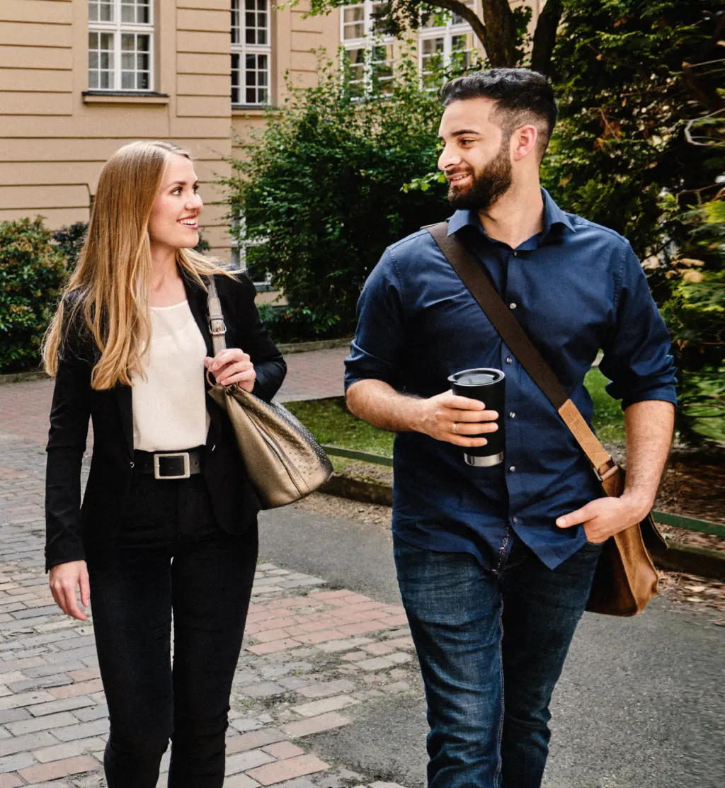 Zwei Personen gehen lächelnd über einen gepflasterten Weg und unterhalten sich. Eine Person trägt eine Tasche über der Schulter, die andere hält einen Becher in der Hand. Im Hintergrund stehen ein historisches Gebäude und Grünflächen.