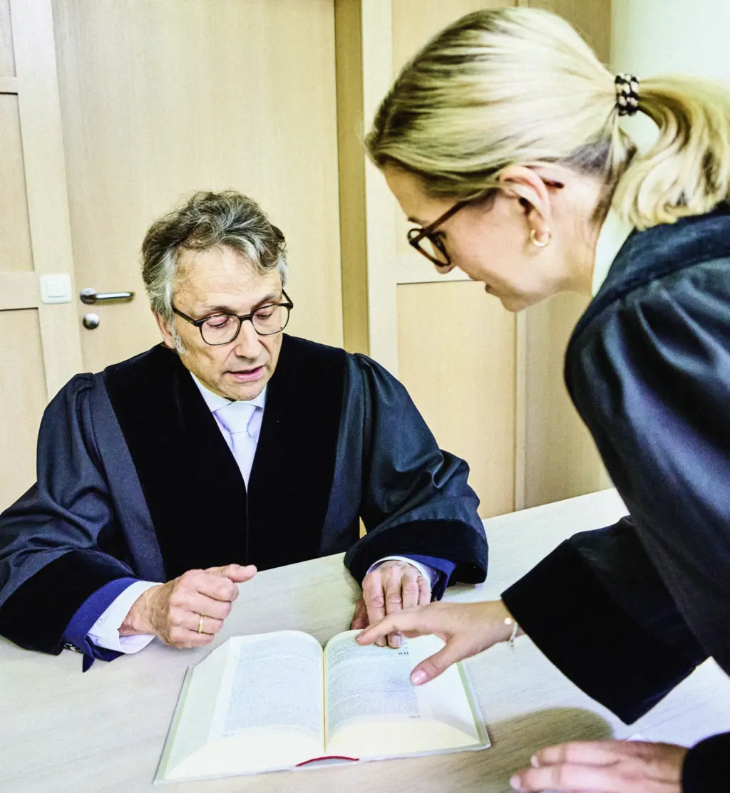 Zwei Personen in Roben stehen bzw. sitzen an einem Tisch und betrachten gemeinsam ein aufgeschlagenes Buch. Eine Person zeigt auf eine Stelle im Buch, während die andere aufmerksam folgt. Die Szene wirkt konzentriert und fachlich.