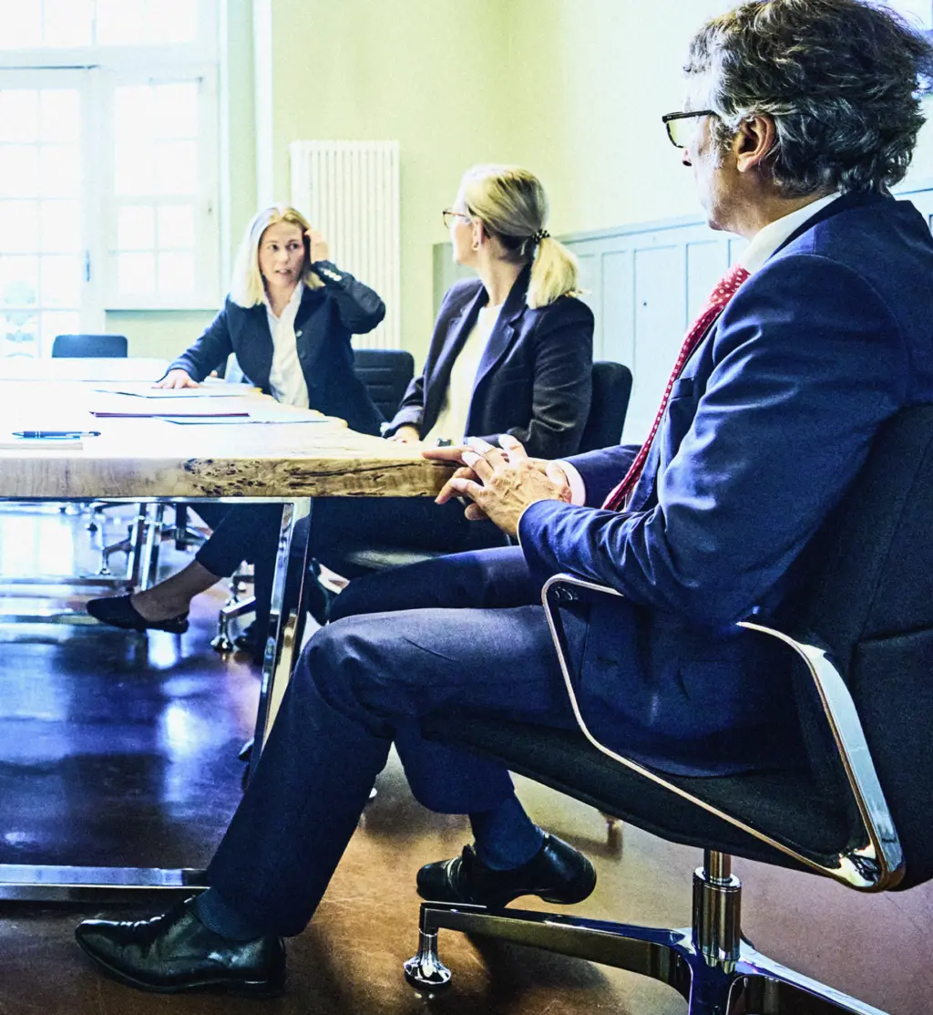 Drei Personen sitzen in einem Besprechungsraum an einem großen Tisch. Zwei Personen auf der gegenüberliegenden Seite sind im Gespräch vertieft, während eine Person im Vordergrund zuhört. Der Raum ist hell und modern eingerichtet, mit großen Fenstern im Hintergrund.