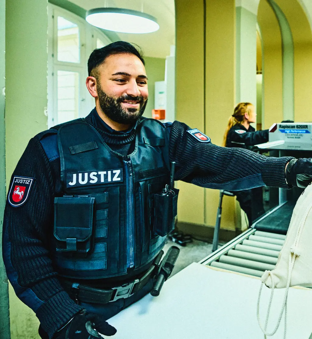 Eine Person in Justizdienstkleidung steht an einer Sicherheitskontrolle und lächelt. Im Vordergrund liegt eine Tasche auf dem Förderband eines Durchleuchtungsgeräts. Im Hintergrund ist eine weitere Person an der Kontrolle tätig. Die Szene wirkt professionell und freundlich.