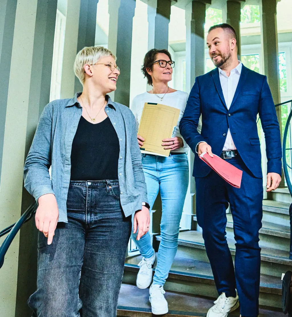 Drei Personen gehen gemeinsam eine Treppe hinunter und unterhalten sich lächelnd. Zwei von ihnen halten Unterlagen in der Hand. Die Umgebung wirkt hell und freundlich, mit großen Fenstern und einem Treppengeländer im Hintergrund.