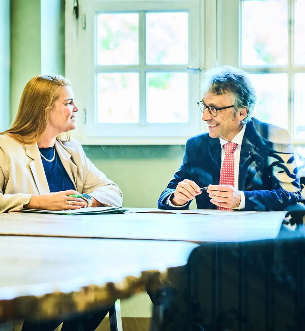 Zwei Personen sitzen einander in einem hellen Besprechungsraum an einem Tisch gegenüber. Eine Frau auf der linken Seite lächelt und hält einen Stift in der Hand. Ein Mann auf der rechten Seite, ebenfalls lächelnd, trägt einen Anzug mit roter Krawatte und hält ein Schreibgerät. Hinter ihnen befindet sich ein großes Fenster, durch das Tageslicht in den Raum fällt.
