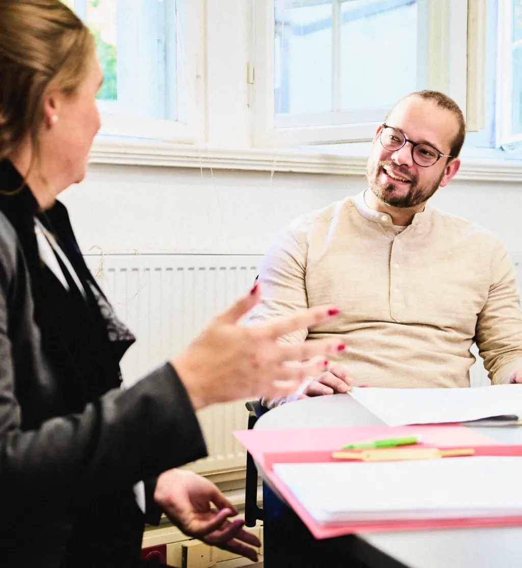 Zwei Personen sitzen in einem hellen Raum an einem Tisch und unterhalten sich. Eine Person gestikuliert, während die andere lächelnd zuhört. Auf dem Tisch liegen Unterlagen und Stifte. Die Atmosphäre wirkt freundlich und kommunikativ.