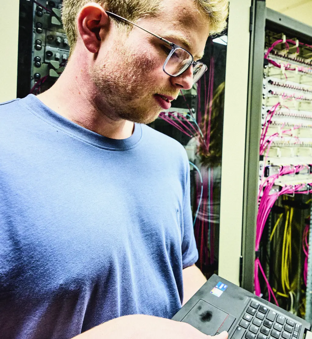 Eine Person steht in einem Technikraum vor einem Serverschrank und arbeitet an einem Laptop. Im Hintergrund sind zahlreiche Kabel und Geräte im Schrank zu sehen. Die Szene wirkt konzentriert und technisch.