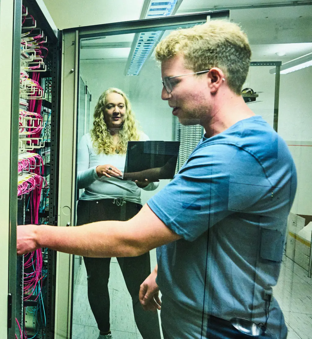 Zwei Personen arbeiten in einem Technikraum an einem Serverschrank mit vielen farbigen Kabeln. Eine Person steht vorne und greift zu einem Kabel, die andere steht dahinter mit einem Laptop in den Händen. Der Raum ist hell beleuchtet und wirkt funktional.