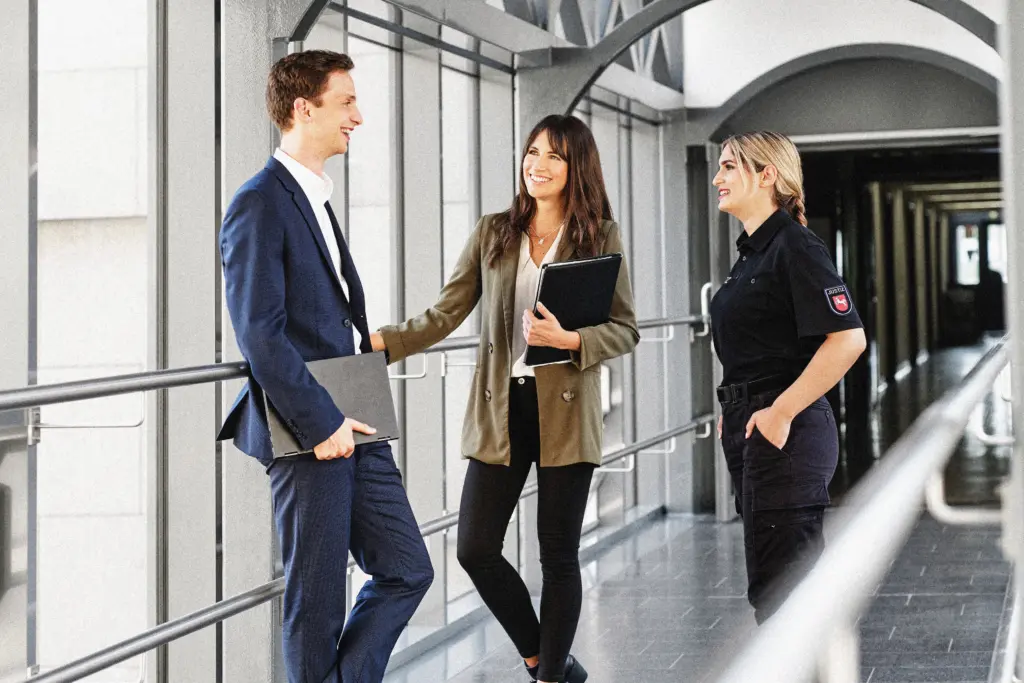 Drei Personen stehen in einem hellen, modernen Gang mit großen Fensterflächen. Eine Frau in Justizuniform spricht mit einem Mann im Anzug und einer Frau in Freizeitkleidung mit einem Laptop im Arm. Alle drei wirken freundlich und im Gespräch vertieft. Die Szene vermittelt einen professionellen und offenen Austausch.
