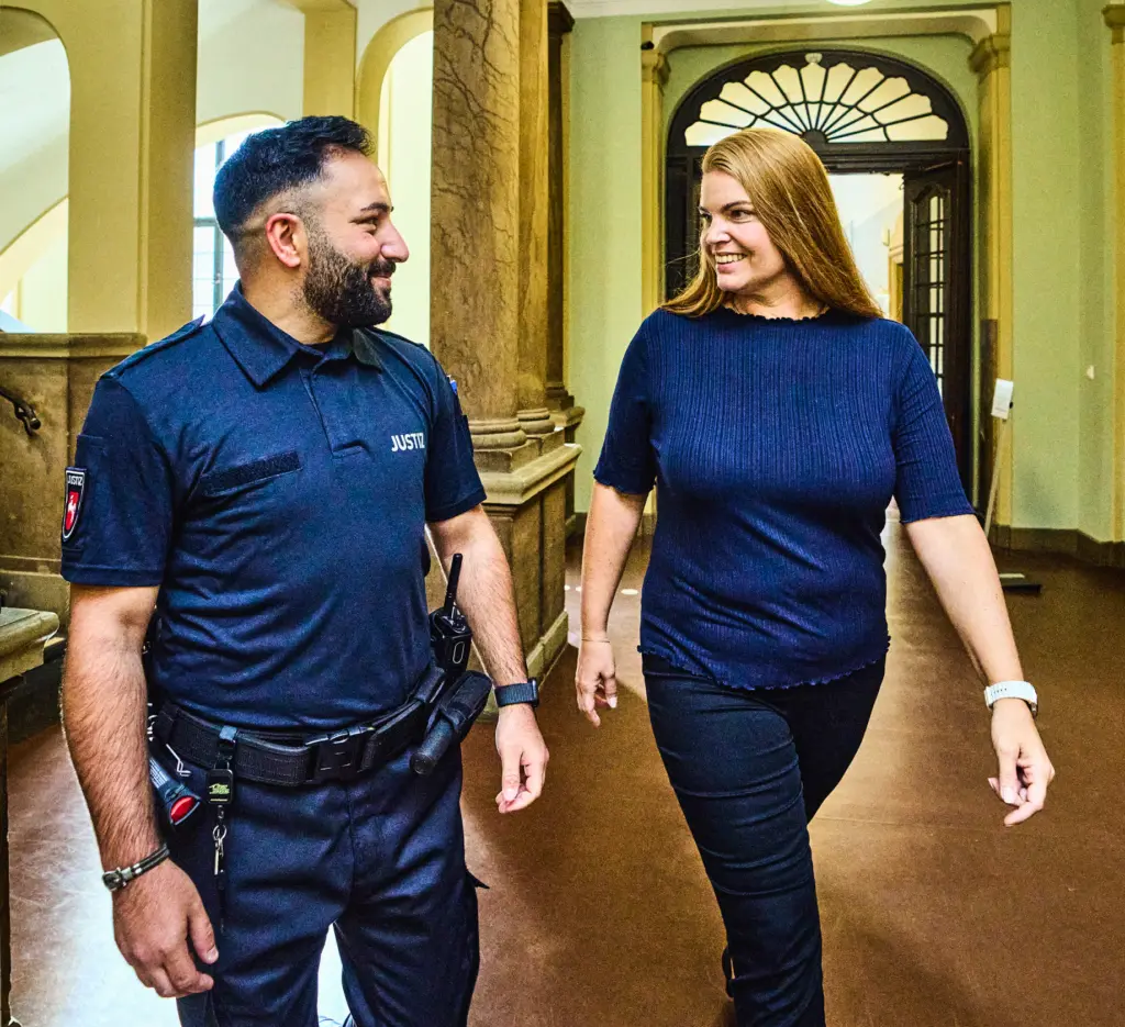 Ein Justizmitarbeiter in dunkelblauer Uniform geht lächelnd einen Flur entlang und unterhält sich mit einer Frau, die neben ihm geht und ebenfalls lächelt. Der Flur wirkt historisch, mit hohen Türen, Rundbögen und Säulen. Im Hintergrund hängen Lampen von der Decke und es steht ein Wasserspender an der Wand. Die Szene vermittelt einen freundlichen und offenen Eindruck.